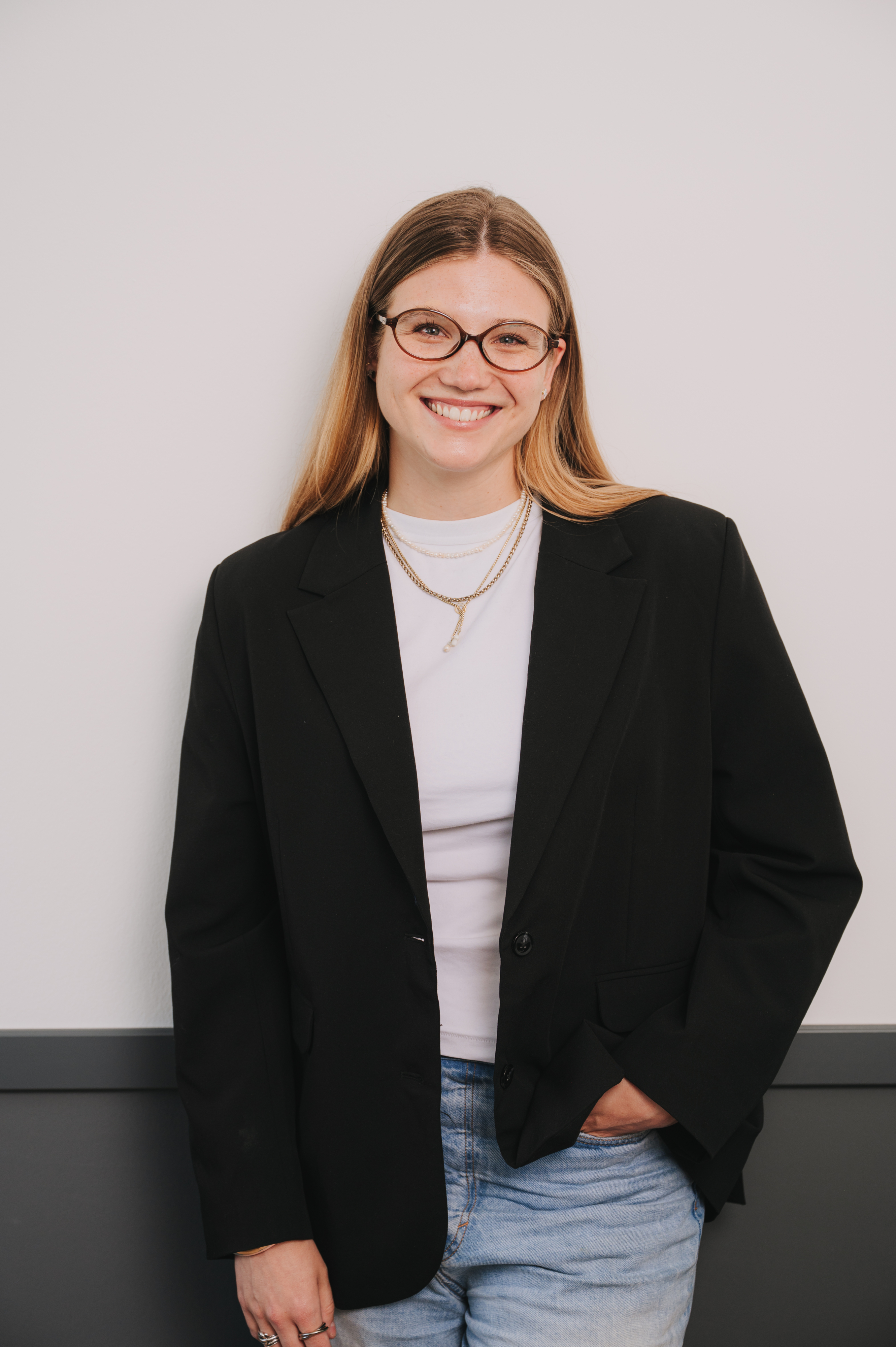 A young woman with blond hair, glasses and wearing a white t-shirt with a black blazer smiling directly at the camera.